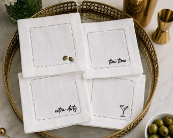 May include: A set of four square white linen cocktail napkins with hemstitched borders. Each napkin features unique black embroidered text or graphics: two green olives, the phrase 'tini time', the phrase 'extra dirty', and a martini glass.