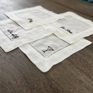 Op de afbeelding: Een set van vier vierkante witte linnen cocktailservetten met zwarte geborduurde tekst en illustraties. De ontwerpen bevatten een martiniglas, de tekst "extra dirty" en de tekst "tea time", omlijst door een decoratieve gestikte rand.