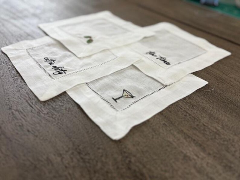 Op de afbeelding: Een set van vier vierkante witte linnen cocktailservetten met zwarte geborduurde tekst en illustraties. De ontwerpen bevatten een martiniglas, de tekst "extra dirty" en de tekst "tea time", omlijst door een decoratieve gestikte rand.