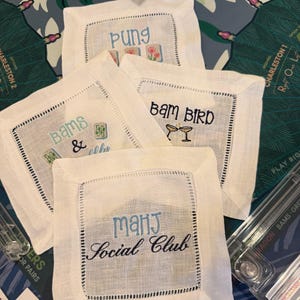 May include: Four square white linen cocktail napkins with hemstitched borders. Each napkin features embroidered text and graphics: one reads Pung with mahjong tiles, one says Bams & with tiles, one says Bam Bird with two glasses, and one says Mahj Social Club.
