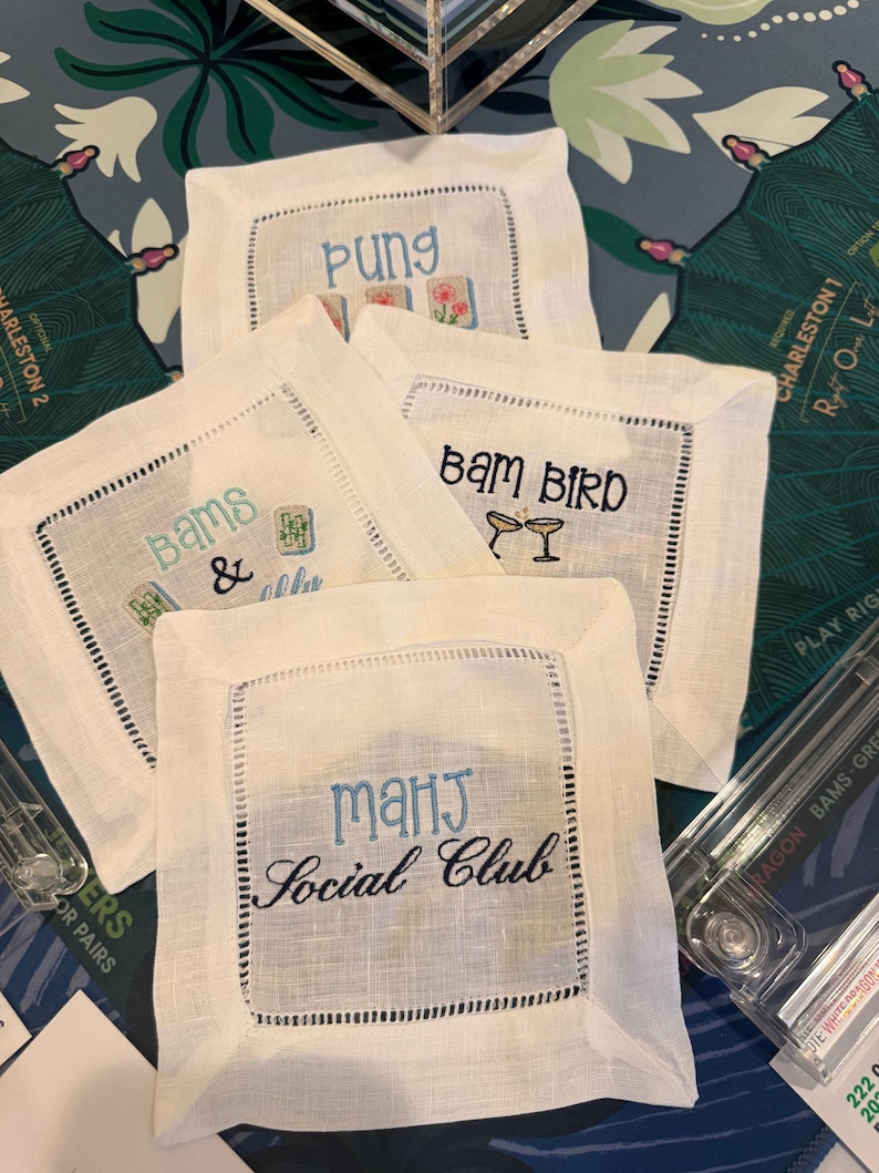 May include: Four square white linen cocktail napkins with hemstitched borders. Each napkin features embroidered text and graphics: one reads Pung with mahjong tiles, one says Bams & with tiles, one says Bam Bird with two glasses, and one says Mahj Social Club.