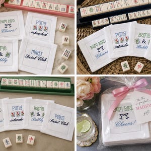 May include: A set of four white square fabric coasters featuring embroidered Mahjong-themed text and graphics including Bam Bird Cheers, Pung intended, Bams & Bubbly, and MahJ Social Club, with some coasters packaged in a clear bag with a pink ribbon.