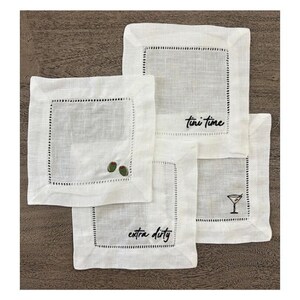 Op de afbeelding: Een set van vier vierkante witte linnen cocktailservetten met ajourzoom. De servetten zijn geborduurd met zwarte tekst Tini time en Extra dirty, samen met kleine groene olijfpictogrammen en een zwarte afbeelding van een martiniglas.
