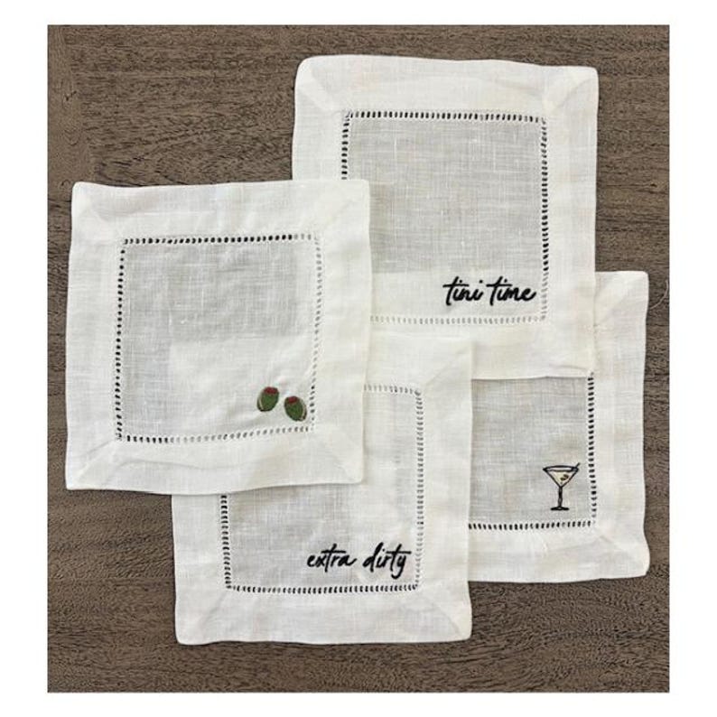 Op de afbeelding: Een set van vier vierkante witte linnen cocktailservetten met ajourzoom. De servetten zijn geborduurd met zwarte tekst Tini time en Extra dirty, samen met kleine groene olijfpictogrammen en een zwarte afbeelding van een martiniglas.