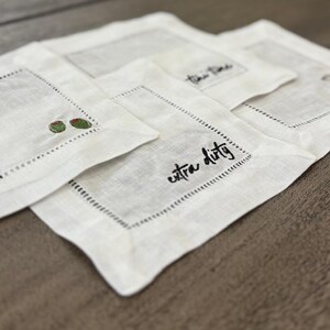 Op de afbeelding: Een set vierkante witte linnen cocktailservetten met ajourzoom. E&eacute;n servet toont de zwart geborduurde tekst extra dirty, terwijl een andere twee kleine groene olijven met rode pimenten in de hoek geborduurd heeft.