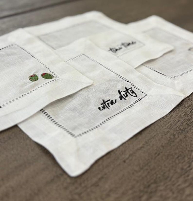 Op de afbeelding: Een set vierkante witte linnen cocktailservetten met ajourzoom. E&eacute;n servet toont de zwart geborduurde tekst extra dirty, terwijl een andere twee kleine groene olijven met rode pimenten in de hoek geborduurd heeft.