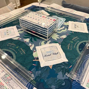 May include: A Mah Jongg set featuring a clear acrylic case filled with white tiles, four clear acrylic tile racks, and a blue patterned game mat. White fabric coasters are present, one embroidered with the text Mahj Social Club.