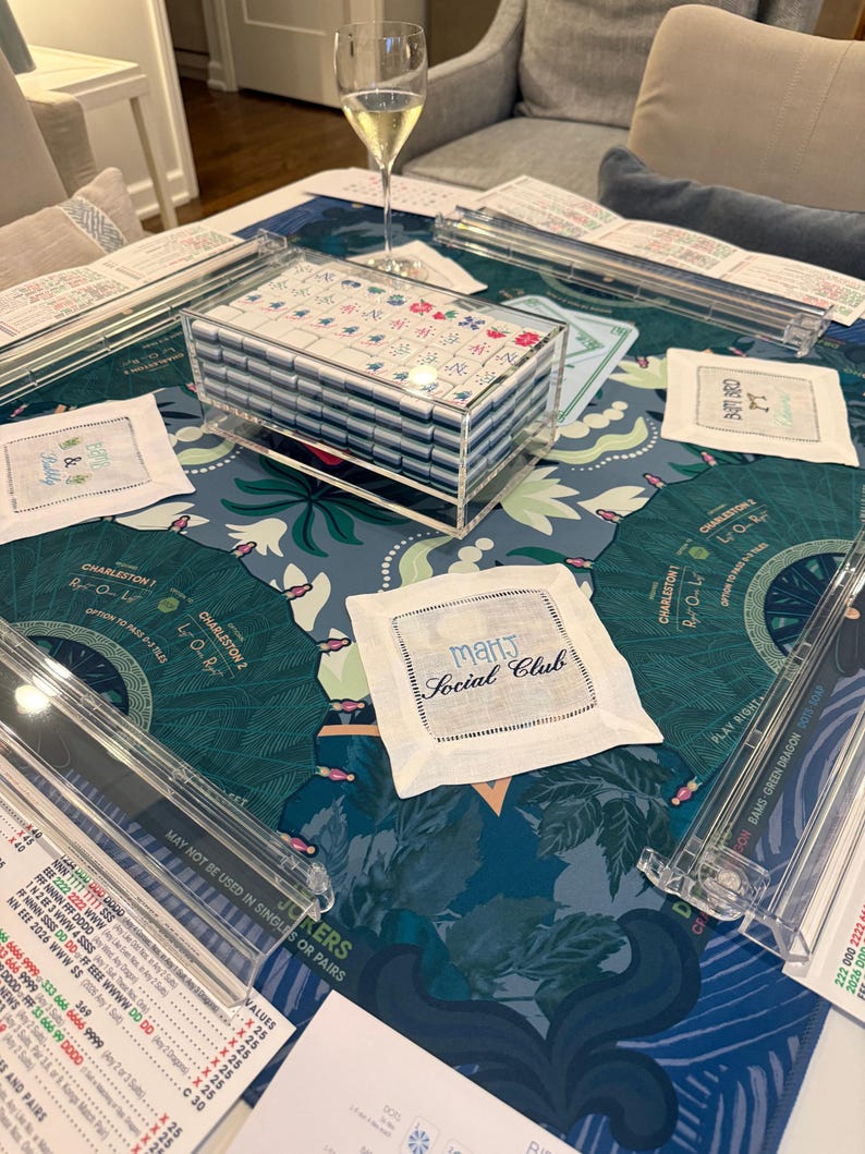 May include: A Mah Jongg set featuring a clear acrylic case filled with white tiles, four clear acrylic tile racks, and a blue patterned game mat. White fabric coasters are present, one embroidered with the text Mahj Social Club.