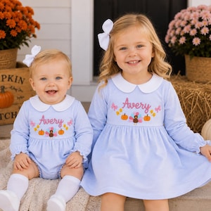 May include: Two light blue and white pinstriped outfits featuring a white Peter Pan collar and long sleeves. The chest area displays embroidered text reading Avery, a central turkey, two orange pumpkins, and small pink bows on each side.