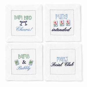 May include: A set of four square white linen coasters with hemstitched borders. Each features colorful embroidery: 'BAM BIRD Cheers!', 'PUNG intended' with three mahjong tiles, 'BAMS & Bubbly' with two mahjong tiles, and 'MAHJ Social Club' in blue and black.