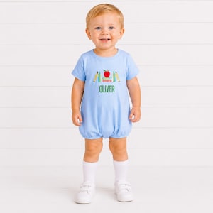 May include: A light blue short-sleeved bubble romper featuring an embroidered graphic of a red apple resting on a stack of books with two yellow pencils. The name OLIVER is embroidered in green capital letters below the school-themed design.