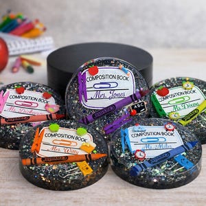 May include: Five round black resin coasters with glitter, each featuring a white label printed with "COMPOSITION BOOK" and a teacher's name, decorated with miniature plastic school supplies including crayons, paper clips, and small apples.