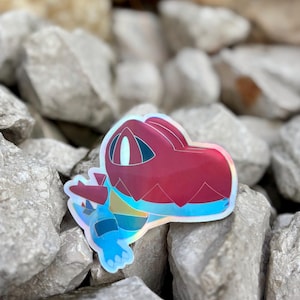 Könnte beinhalten: Ein holografischer Aufkleber mit einer cartoonartigen, krokodilartigen Kreatur. Der Aufkleber hat einen roten Kopf, einen blauen Körper und schillernde Akzente. Der Aufkleber liegt auf einem Bett aus grauen Steinen.