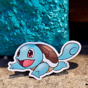 Puede incluir: Una pegatina troquelada con el personaje de dibujos animados Squirtle. La pegatina tiene un cuerpo azul claro, un caparazón marrón y una cara sonriente con ojos rojos y boca rosa. La pegatina está sobre una superficie texturizada.