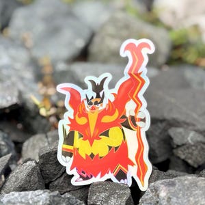 Puede incluir: Una pegatina holográfica con un personaje de dibujos animados estilizado con un diseño de fuego. El personaje tiene un esquema de color rojo y amarillo con acentos negros. La pegatina está colocada sobre un lecho de rocas de color gris oscuro.