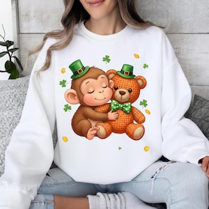 Può includere: Felpa bianca con un design per la festa di San Patrizio. La grafica mostra una scimmia marrone che abbraccia un orsacchiotto arancione, entrambi con cappelli e papillon verdi. Monete d'oro e trifogli verdi sono sparsi.