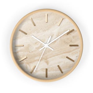 Puede incluir: Un reloj de pared redondo con marco de madera clara y esfera con patrón de mármol beige. El reloj cuenta con doce marcadores de hora rectangulares de color oro metálico y tres manecillas blancas que indican la hora sobre la textura.