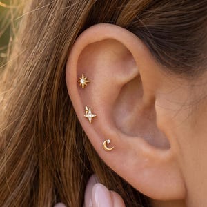 Puede incluir: Tres pendientes de botón dorados dispuestos a lo largo del cartílago y el lóbulo de una oreja. El pendiente superior es una estrella de cuatro puntas, el central tiene forma de estrella radiante y el inferior es una pequeña luna creciente.