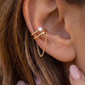 May include: A gold-toned ear cuff featuring two parallel bands connected by a delicate chain. The upper band is accented with a single small, clear, round-cut stone. The piece is designed to wrap around the cartilage of the ear, creating a layered look.