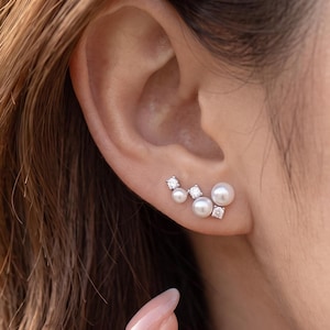 May include: An ear climber earring featuring a curved design with three white round pearls and two clear faceted stones set in a polished silver-tone metal base, following the natural contour of the earlobe along the outer edge.