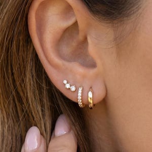 May include: A set of three gold earrings worn in the earlobe and cartilage. The set includes a small plain gold hoop, a small gold hoop featuring a row of clear stones, and a small gold stud earring with three clear stones in a cluster.