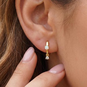 Op de afbeelding: Een gouden oorring met een kleine, ronde, witte opaalsteen in de ringband en een enkele, piepkleine, heldere, rondgeslepen edelsteen die aan de onderkant van de ring hangt, wat zorgt voor een verfijnde uitstraling aan de oorlel.