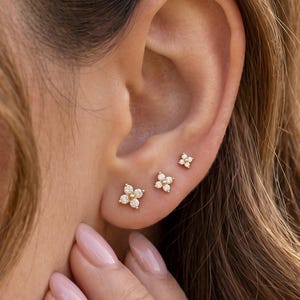 May include: Three gold stud earrings shaped like four-petaled flowers, each featuring small clear stones. The earrings are arranged in a row along the earlobe, with the largest flower at the bottom and two smaller ones positioned above it.