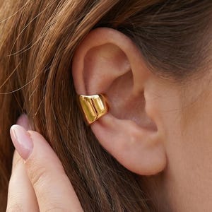 May include: A polished gold-tone ear cuff with a smooth, wide, and slightly curved band design. The jewellery piece sits securely on the cartilage of the ear, featuring a reflective metallic surface that catches the light with its simple, solid form.