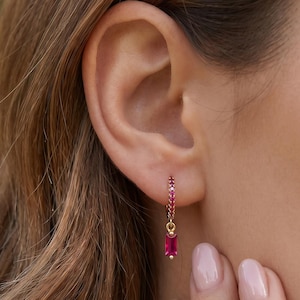 May include: A gold hoop earring featuring a row of small, vibrant pink stones along the hoop and a single, rectangular, baguette-cut pink stone dangling from the bottom. The earring is crafted with a polished gold finish and a secure clasp mechanism.