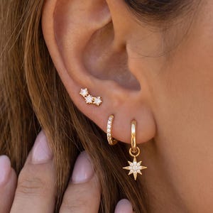 May include: A set of gold-toned earrings consisting of a small stud featuring three tiny clear stones arranged in a cluster, a plain gold hoop, and a gold hoop earring with a dangling eight-pointed star charm encrusted with small clear stones.