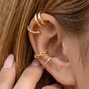 May include: A set of four gold-toned ear cuffs worn on the cartilage and conch. The collection includes a smooth double-band cuff, a beaded texture cuff, a small chain-link cuff, and a simple thin band cuff, all featuring a polished metallic finish.