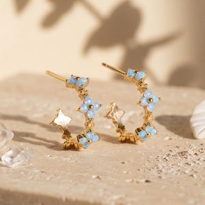 May include: A pair of gold hoop earrings featuring small, light blue, flower-shaped stones arranged along the outer edge. The earrings have a post-back closure and are displayed on a textured, light-colored stone surface.