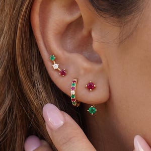 May include: A set of gold earrings featuring a curved climber with green, white, and red stones, a small hoop with alternating green and white stones, a single red stud, and a single green stud, all worn in multiple piercings on one ear.