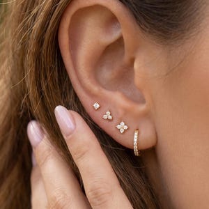 Peut inclure: Un ensemble de quatre boucles d'oreilles en or comprenant trois petites puces d'oreilles et une petite créole. Les puces présentent des pierres claires en arrangements simples, triples et quadruples, tandis que la créole est ornée de petites pierres.
