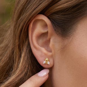 May include: A gold stud earring shaped like a small bee, featuring a metallic gold body and two clear, marquise-cut stones serving as wings. The earring is secured to the earlobe, showcasing a detailed insect design with a polished finish.