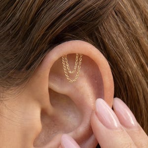 May include: A gold-toned ear cartilage piercing featuring two delicate, interconnected fine chains that drape in a soft curve along the upper ear rim. The jewellery is secured at two points along the cartilage, creating a layered, dangling effect.