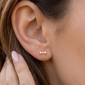 May include: A gold stud earring featuring three small, clear, round-cut stones arranged in a horizontal row, with a delicate gold chain draped in a U-shape connecting the two outer stones, creating a subtle, dangling effect on the earlobe.