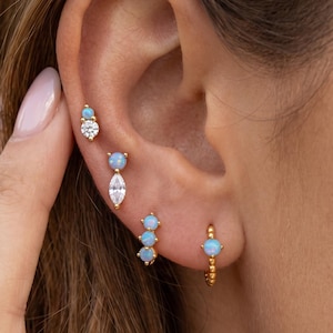 Peut inclure: Un ensemble de boucles d'oreilles dorées ornées de cabochons bleu clair style opale et de pierres transparentes taille marquise et ronde. La collection comprend trois clous d'oreilles aux compositions variées et une petite créole perlée.