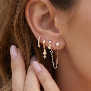 May include: A collection of gold earrings, including a small hoop with clear stones, a small hoop with a dangling diamond-shaped charm, and a pearl stud with a chain. The earrings are worn on the ear.