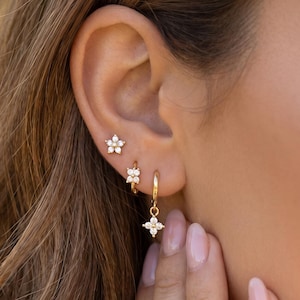 May include: A set of three gold-toned earrings featuring flower-shaped designs with clear, sparkling stones. The top earring is a stud, the middle is a small hoop, and the bottom is a hoop earring with a dangling flower-shaped charm.