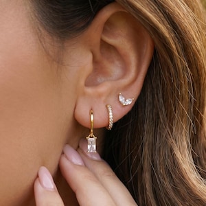 Könnte beinhalten: Nahaufnahme eines Ohrs mit mehreren goldenen Ohrringen. Das Ohr zeigt einen kleinen Creolen-Ohrring mit einem rechteckigen Edelstein, einen kleinen Creolen-Ohrring mit winzigen Edelsteinen und einen Ohrstecker mit Schmetterlingsdesign.