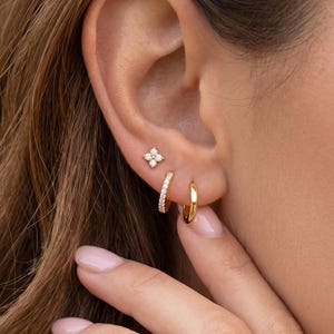 Peut inclure: Un ensemble de trois boucles d'oreilles en or portées à une oreille. La boucle supérieure est un petit clou en forme de fleur à quatre pétales avec des pierres claires. La boucle centrale est un petit anneau orné de pierres claires.