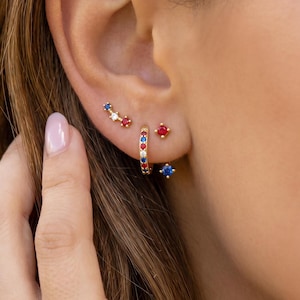 Könnte beinhalten: Eine Auswahl an goldfarbenen Ohrringen mit roten, weißen und blauen Edelsteinen. Die Ohrringe umfassen einen kleinen Creolen, einen Stecker und einen Kletterstil. Der Schmuck wird an einem Ohr präsentiert.