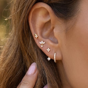 Puede incluir: Primer plano de una oreja adornada con múltiples pendientes dorados. Los pendientes incluyen un aro pequeño, un pendiente con una sola gema, un pendiente cuadrado pequeño, un diseño floral y un pendiente en forma de estrella. Los pendientes están engastados con pequeñas gemas transparentes.