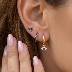 May include: A close-up of three earrings on an ear. The earrings include a gold stud with a blue stone, a gold hoop with blue stones, and a gold hoop with an eye-shaped charm with blue stones.