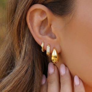 May include: A set of three gold-toned earrings worn in a single ear. The set includes a small twisted hoop, a rectangular hoop featuring a row of clear stones, and a smooth, polished teardrop-shaped hoop earring with a reflective gold finish.