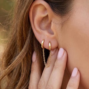 Könnte beinhalten: Ein goldener Doppel-Creolen-Ohrring, der durch eine zarte, feine Kette verbunden ist. Das Schmuckstück besteht aus zwei kleinen, polierten Goldringen für zwei separate Ohrlöcher, die durch eine dünne, glänzende Goldkette verbunden sind.
