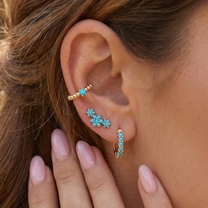 Puede incluir: Un ear cuff dorado con una piedra turquesa, un pendiente de botón dorado con tres grupos de flores de turquesa y un pequeño aro dorado con varias piedras turquesas incrustadas a lo largo del borde frontal del aro.