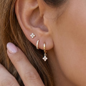 Puede incluir: Primer plano de una oreja adornada con tres pendientes dorados. Incluye un pequeño pendiente de botón con diseño floral, un aro pequeño con piedras transparentes y un aro con un charm floral colgante. Fondo borroso.