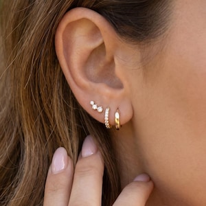 May include: A woman's ear is shown with three gold earrings. There is a small hoop, a curved bar with small clear stones, and a stud design with three clear stones. The earrings are set against the backdrop of the woman's brown hair.